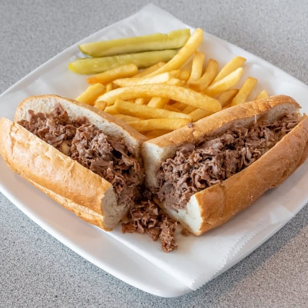 Cheesesteak sandwich cut in half, served with golden french fries and pickle spears on a white plate.